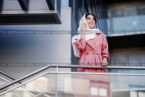 Young Muslim Woman walking on the street wearing hijab headscarf using smart phone in a office building. Business background.. Young Muslim woman wearing hijab using her smartphone. Стоковое фото, агентство Ingram Publishing / Фотобанк Лори