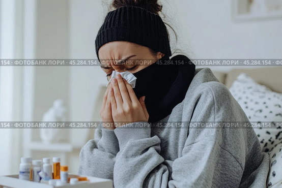 Young sick woman with cold or flue sitting on grey couch, holding a tissue to clear, blowing runny nose. Sneezing Girl under gray plaid blanket with handkerchief to dripping nose, blowing snot at home. Стоковое AI-изображение, AI-художник Марина Демидюк / Фотобанк Лори