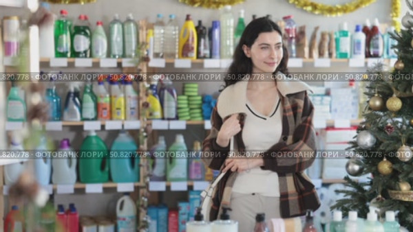 Young Spanish woman chooses household chemicals in a supermarket decorated for Christmas. Girl in a coat came to the hardware store on New Year's Eve. Стоковое видео, видеограф Яков Филимонов / Фотобанк Лори