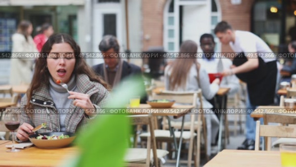 Young woman in a jacket sits on a cool terrace of a restaurant and eats a salad against the background of other visitors. European woman tries a freshly prepared vegetable salad from a chef. Стоковое видео, видеограф Яков Филимонов / Фотобанк Лори