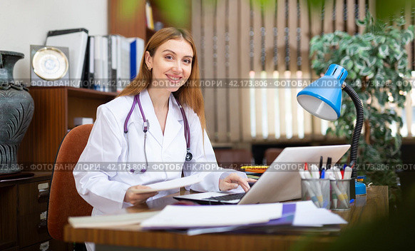 Young woman internist studies the patient's outpatient card, typing the treatment appointment on the computer in the office. Стоковое фото, фотограф Яков Филимонов / Фотобанк Лори