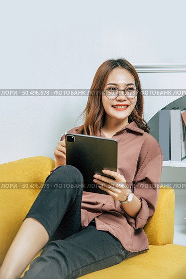 Young woman wearing glasses sitting on sofa and using technology of digital pen on tablet to working from home during self-isolation and quarantine covid-19 coronavirus outbreak. Стоковое фото, агентство Ingram Publishing / Фотобанк Лори