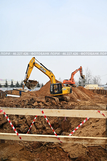 Желтый экскаватор JCB и оранжевый экскаватор ведут земляные работы на строительной площадке, разрабатывая котлован. В переднем плане видно деревянное ограждение с красно-белой лентой, траншея и строительные материалы. (2024 год). Редакционное фото, фотограф Александр Орымбаев / Фотобанк Лори