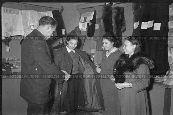 Женщина меряет пальто рядом с прилавками ярмарки, покупательницы примеряют верхнюю одежду перед покупкой, торговые витрины на заднем плане, Якутия, январь 1969 год. Редакционное фото, фотограф Варфоломей Тетерин / Фотобанк Лори